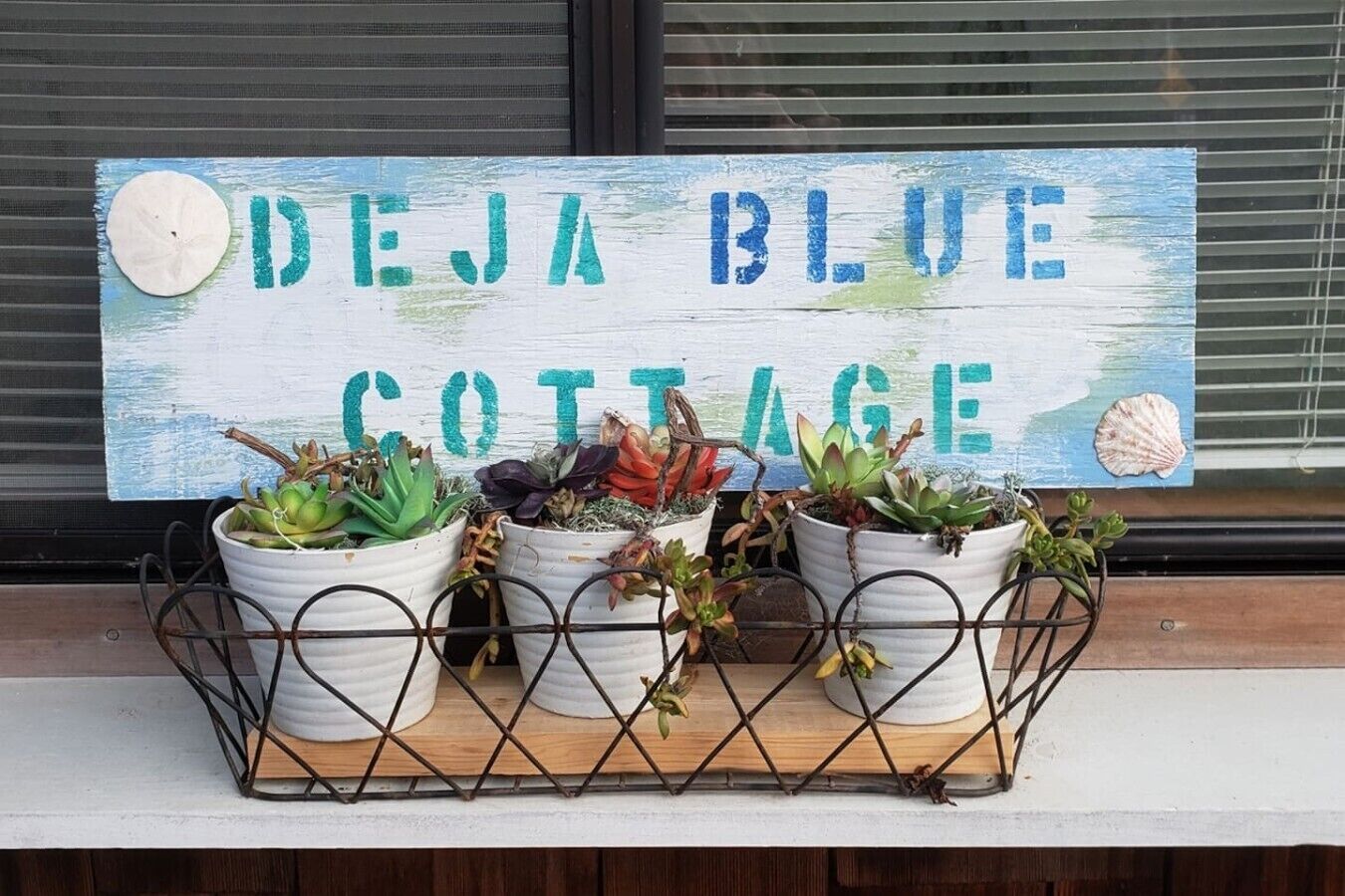 Un cartel con 'DEJA BLUE COTTAGE' y tres plantas en macetas frente a una ventana.