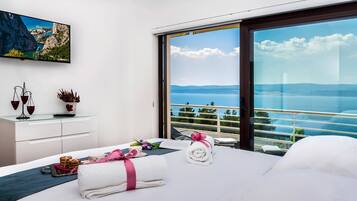 A modern hotel room with a large bed, a bedside table with a vase and a scale, a view of the sea through floor-to-ceiling windows, and a painting above the bed.