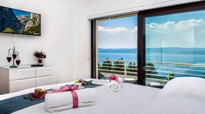 Habitación de hotel moderna con una cama grande, una mesita de noche con un florero y una balanza, vistas al mar a través de ventanales del suelo al techo, y un cuadro sobre la cama.