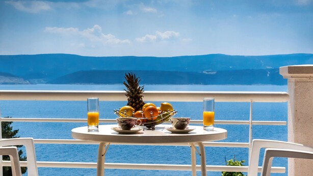 Ein Tisch mit Obstschale, zwei Orangensaftgläsern und einer Tasse auf einem Balkon mit Blick auf ein Gewässer und Berge.