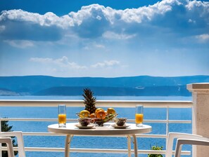 Una mesa con frutero, dos vasos de jugo de naranja y una taza en un balcón con vista al agua y montañas.