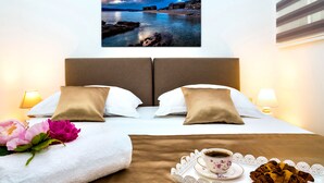 Ein ordentlich bezogenes Bett mit weißen und beige farbenen Kissen, ein Tablett mit einer Tasse Kaffee und einem Croissant sowie ein Blumenstrauß.