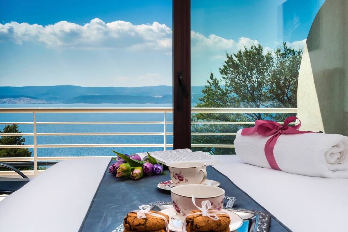 A balcony with a view of the sea and mountains, a tray with cookies and a teapot, and a neatly made bed with a towel.