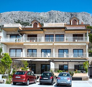 Un edificio de tres pisos con balcones, fachada de piedra y montañas al fondo.