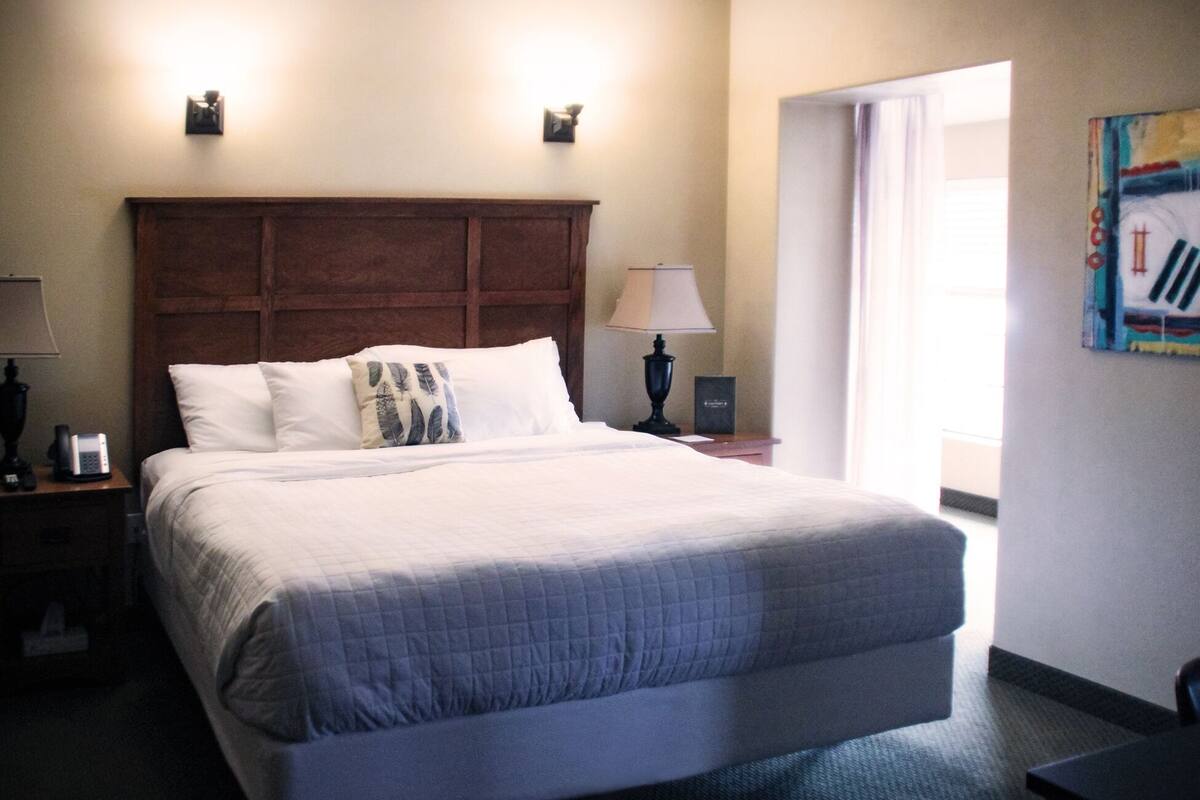 A hotel room with a large bed, a nightstand with a lamp, a bedside table, and a door leading to a bright area.