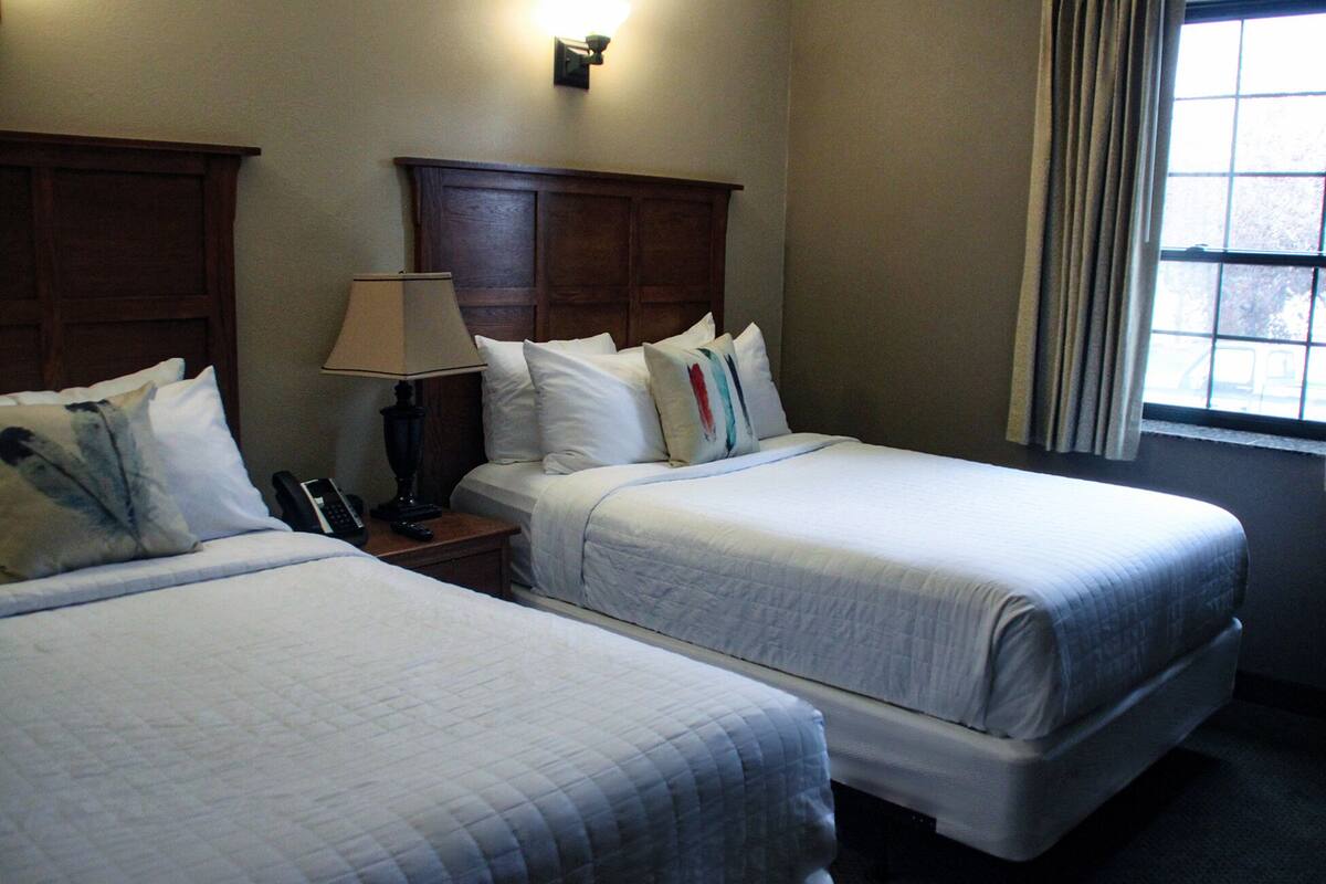 A hotel room with two single beds, a nightstand with a lamp, a telephone, and a window with curtains.