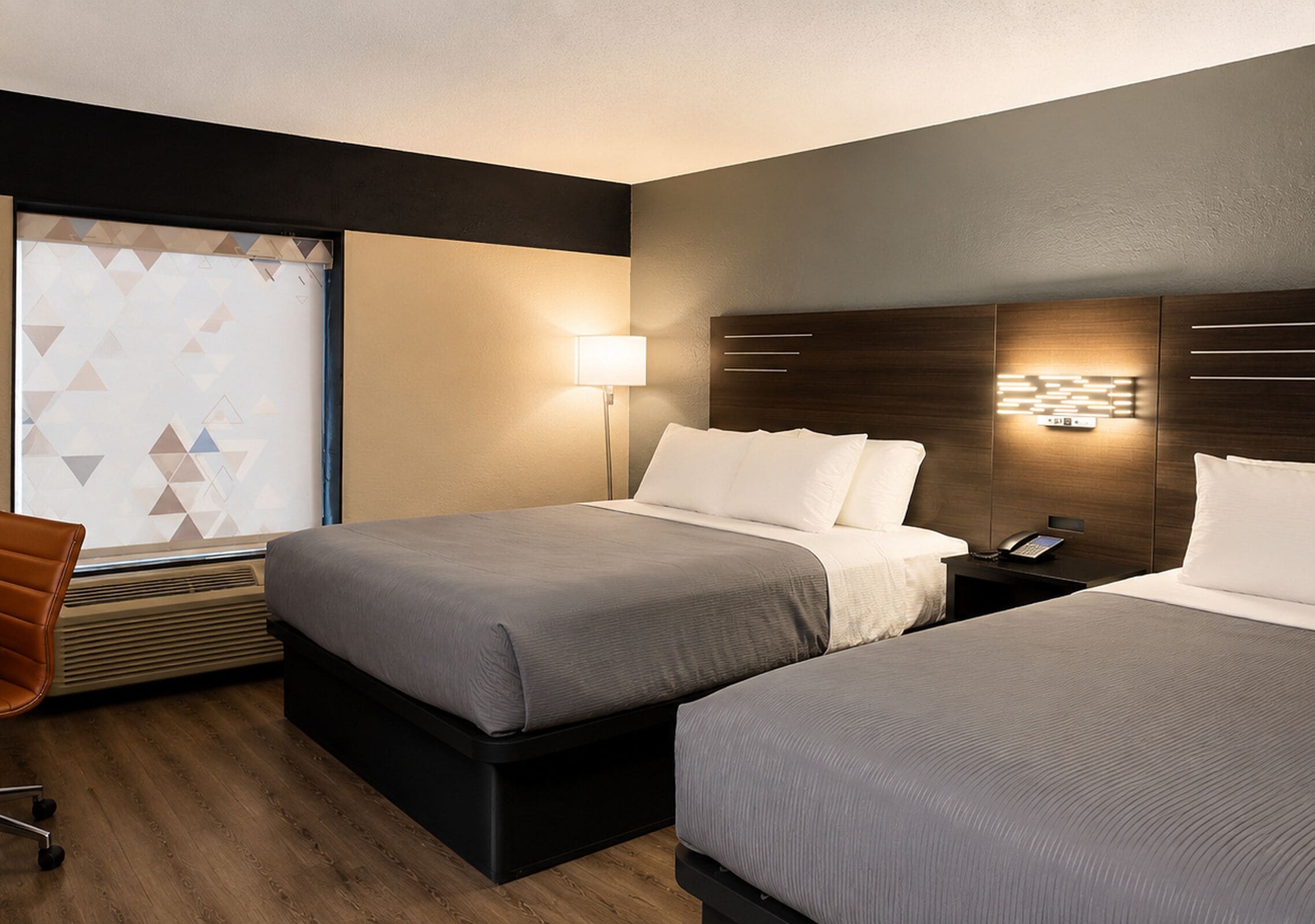 Habitación de hotel con dos camas, un escritorio, una silla, un ventanal con persianas geométricas y una lámpara fijada a la pared.