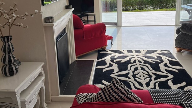A living room with a red sofa, a black and white patterned rug, a fireplace, and a view of the sea through large glass doors.