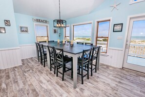 A dining area with a large table and chairs, a hanging light fixture, and a view of the beach through large windows.