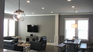 A spacious living room with a flat-screen TV, comfortable seating, a dining area, and large windows.