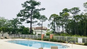 A swimming pool with lounge chairs and a table, surrounded by a white fence and tall trees.