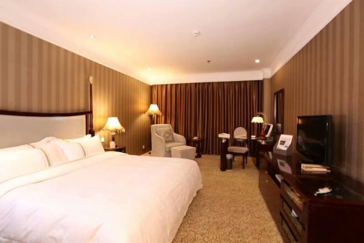 A hotel room with a large bed, two armchairs, a desk with a TV, and a wall-mounted TV.
