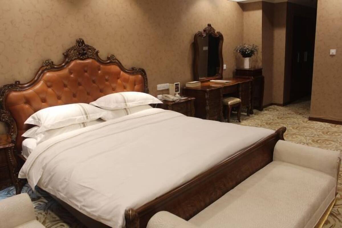 A hotel room with a large bed, a wooden headboard, a desk with a chair, a mirror, and a vase with flowers.