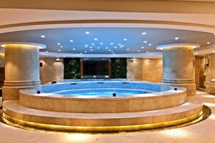 Una bañera de hidromasaje circular con suelo de mármol, rodeada de columnas y un techo empotrado con luces.