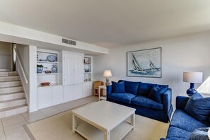 Un salón con un sofá azul, una mesa de centro blanca y un cuadro de veleros en la pared.