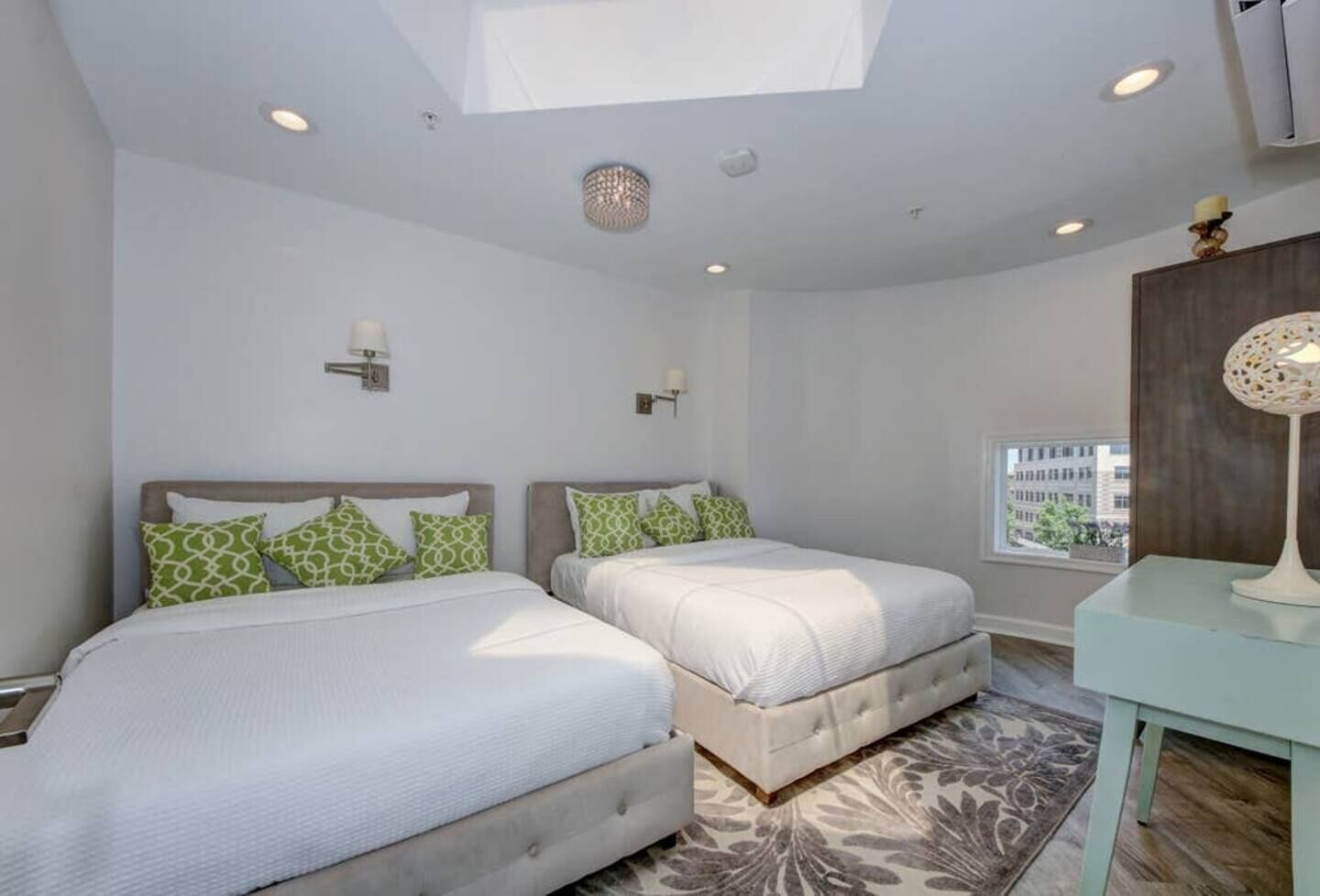 A modern bedroom with two beds, a skylight, recessed lighting, and a view of the city.