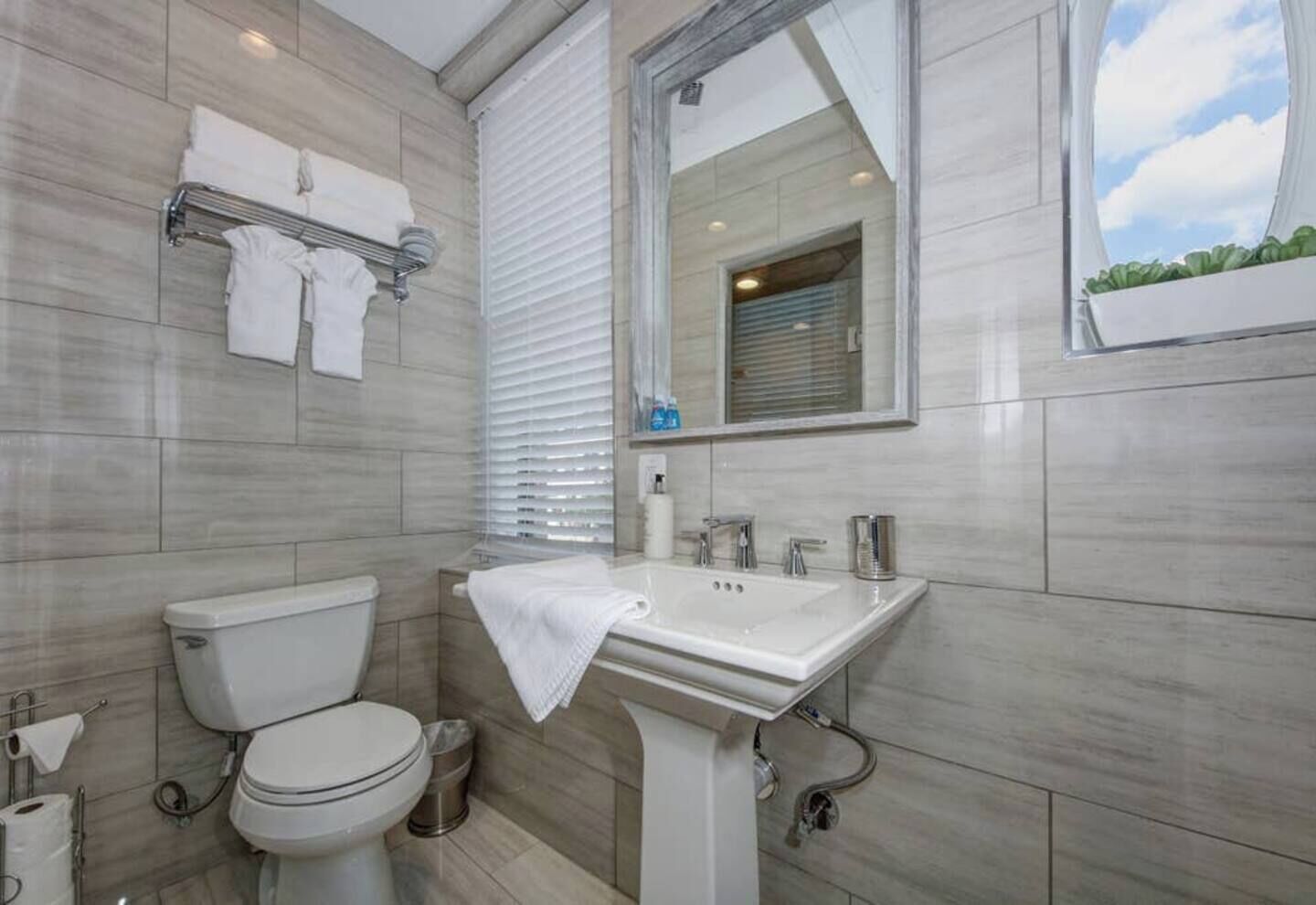A modern bathroom with a toilet, sink, and mirror. There are towels on a rack and a window with blinds.