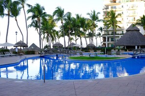 Piscina com espreguiçadeiras e guarda-sóis de palha, cercada por palmeiras e um edifício de vários andares.