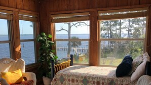 A cozy room with wicker furniture, a bed with a floral bedspread, and large windows overlooking a body of water. - Amazing Ochlockonee Riverfront Home!  (Panacea)
