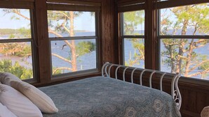 A bedroom with a large bed, wooden walls, and a view of a lake and trees through two windows. - Amazing Ochlockonee Riverfront Home!  (Panacea)