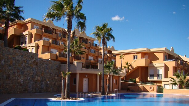 A swimming pool with a stone wall and palm trees, surrounded by multi-story buildings with balconies. - Beautiful apartment with views (Pedreguer)