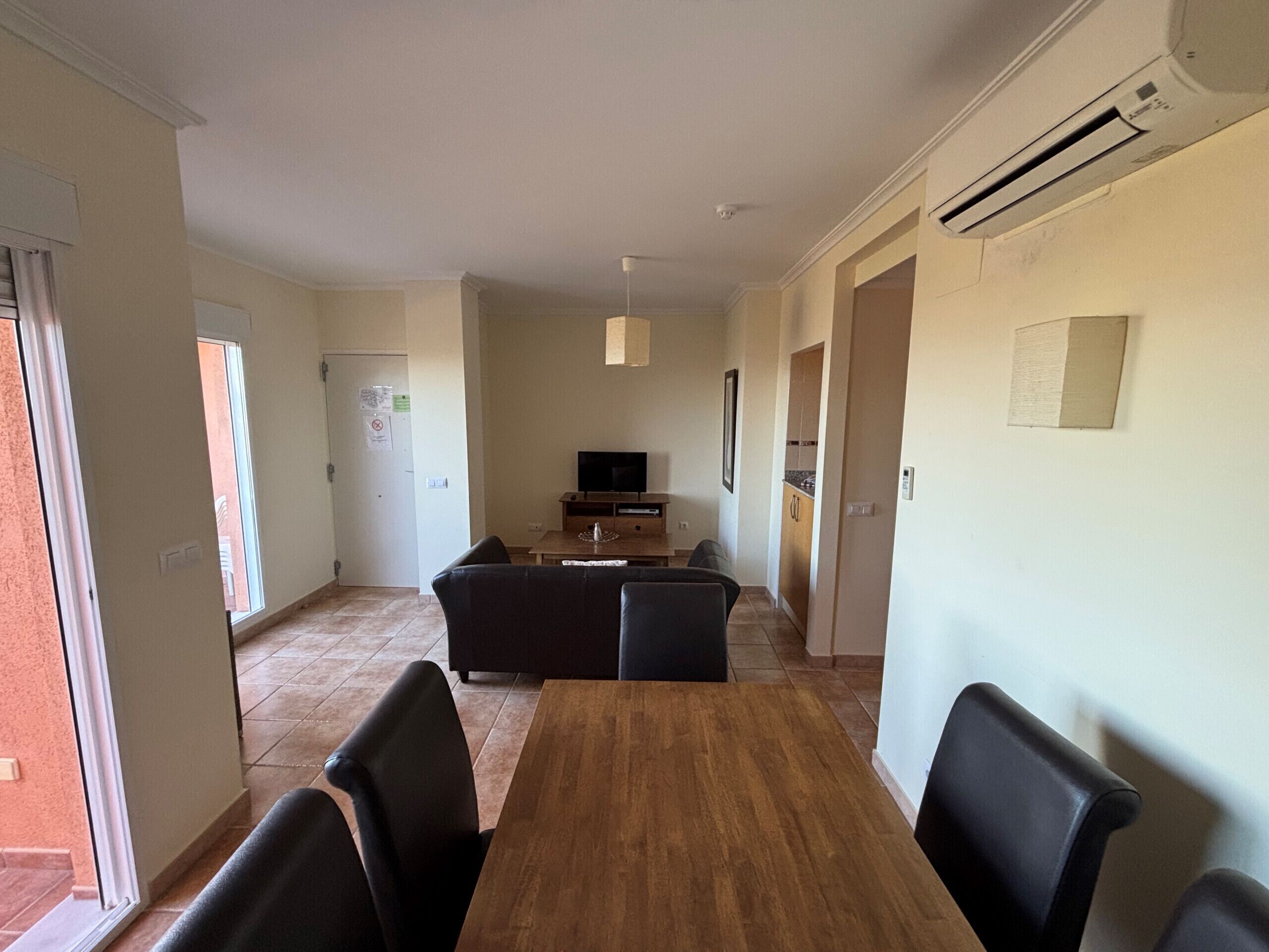 Un comedor con una mesa de madera y sillas negras, un televisor al fondo y una cocina visible a través de una puerta abierta.