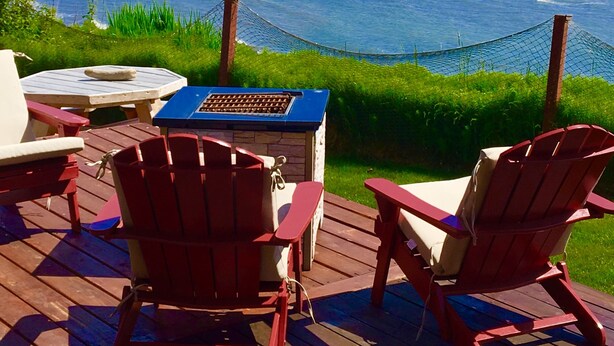 Eine Terrasse mit roten Adirondack-Stühlen, einem Tisch und einem Grill mit Blick auf ein Gewässer, im Hintergrund Berge.