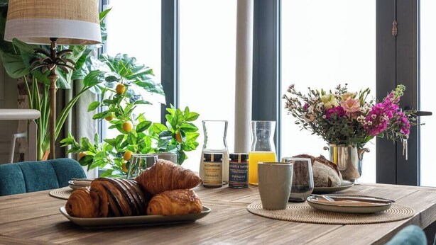 Une table de petit-déjeuner avec des croissants, un vase de fleurs et une bouteille de jus d’orange.