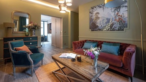 Un salon avec un canapé en velours rouge, un fauteuil vert, une table basse en bois et un grand tableau accroché au mur.
