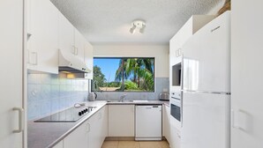 Een compacte keuken met witte kastjes, een vaatwasser en een raam met uitzicht op palmbomen.