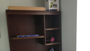 A small, compact workspace with a wooden desk, a black office chair, a shelf with a book, a pink hairdryer, and a box on top. - ENCANTO CERCA DEL AEROPUERTO - APARTAESTUDIO PEQUEÑO (Bogota)