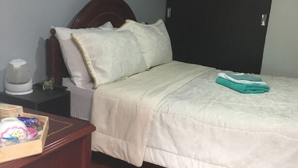 A bedroom with a wooden bed, white bedding, a nightstand with a kettle, and a painting on the wall. - ENCANTO CERCA DEL AEROPUERTO - APARTAESTUDIO PEQUEÑO (Bogota)