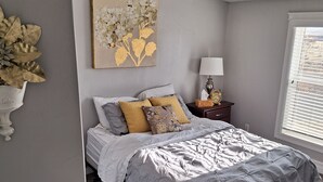 Ein Schlafzimmer mit Bett, Nachttisch mit Lampe, Wanduhr und Fenster mit Jalousien.