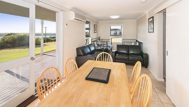 A spacious living area with a wooden dining table, two black leather sofas, and a glass door leading to a balcony.