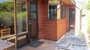 Una cabaña de madera con puerta corredera de vidrio, un patio con mesa y sillas, y una parrilla para barbacoa.