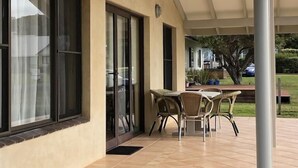 Un patio cubierto con mesa y sillas, ventanas amplias y vistas a un jardín.