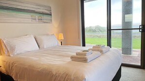 A hotel room with a large bed, white linens, a painting on the wall, and a view of the outdoors through a glass door.