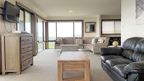 A spacious living room with a large window, a leather sofa, a wooden coffee table, and a wooden cabinet.
