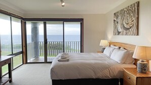 A hotel room with a large bed, two bedside tables with lamps, a painting on the wall, and a balcony with a view of the sea.