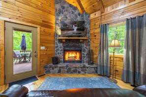 A log cabin interior with a stone fireplace, wooden walls, a door leading outside, and a window with curtains.