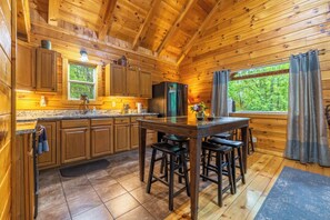 A wooden cabin interior with a kitchen area, wooden cabinets, a refrigerator, a dining table with stools, and a window with curtains.