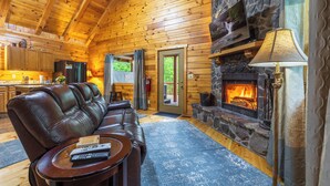 A log cabin interior with a stone fireplace, leather sofa, and a television mounted above it. - Hot Tub, Fire Pit, Large Decks & Blackstone Griddle (Logan)