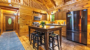 A wooden cabin interior with a kitchen featuring a stainless steel refrigerator, wooden bar stools, and a wooden dining table. - Hot Tub, Fire Pit, Large Decks & Blackstone Griddle (Logan)