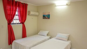 A room with two single beds, a window with red curtains, an air conditioning unit, and a painting on the wall. - La Bienvenue Rentals (Willemstad)