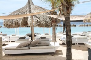 Un espace de détente aménagé sur la plage, avec des chaises longues blanches et des parasols en chaume, offrant une vue sur une mer calme.