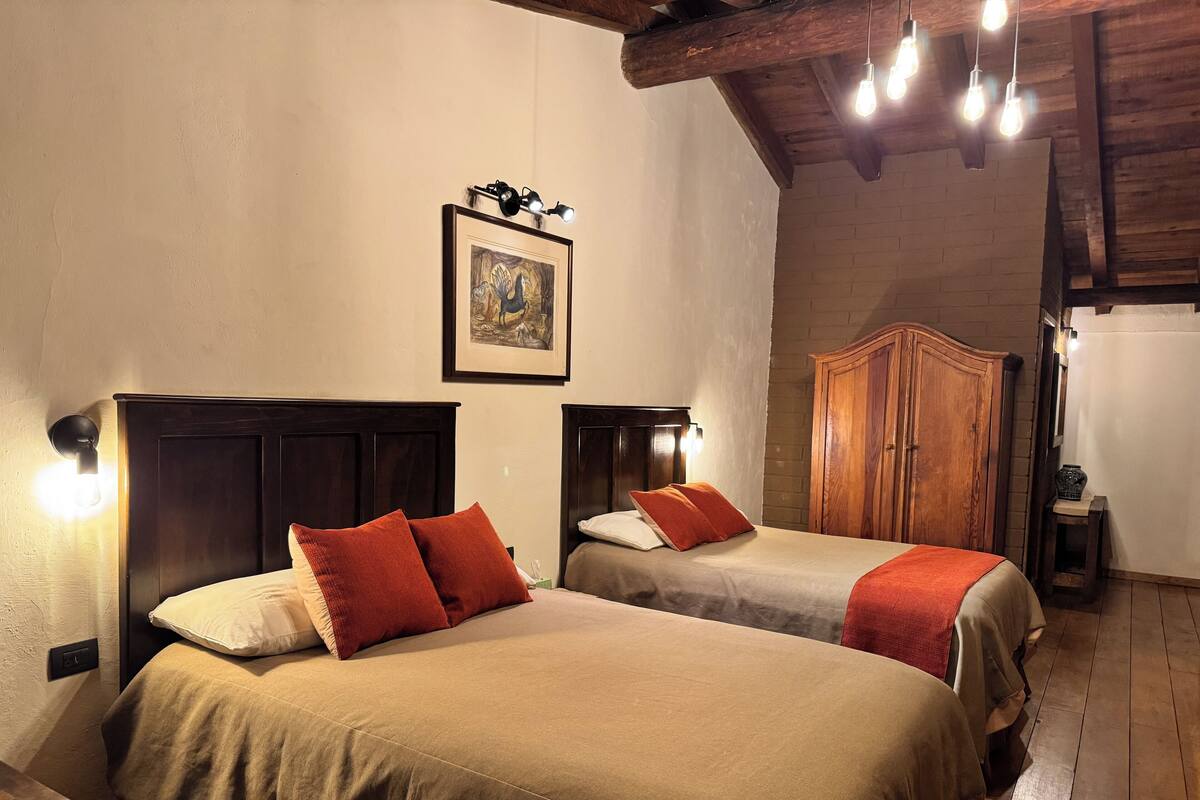 Una habitación con dos camas, cada una con cabecero y lámparas de noche. Hay un armario de madera y un cuadro en la pared.