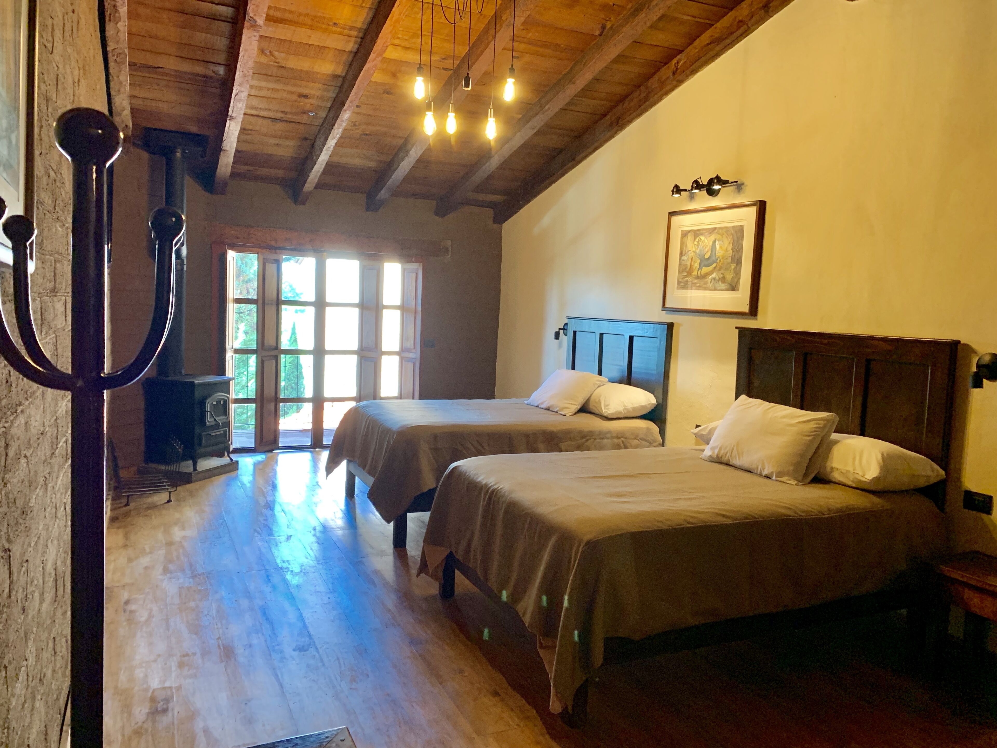 A room with two beds, wooden floors, a large window, and a rustic ceiling.