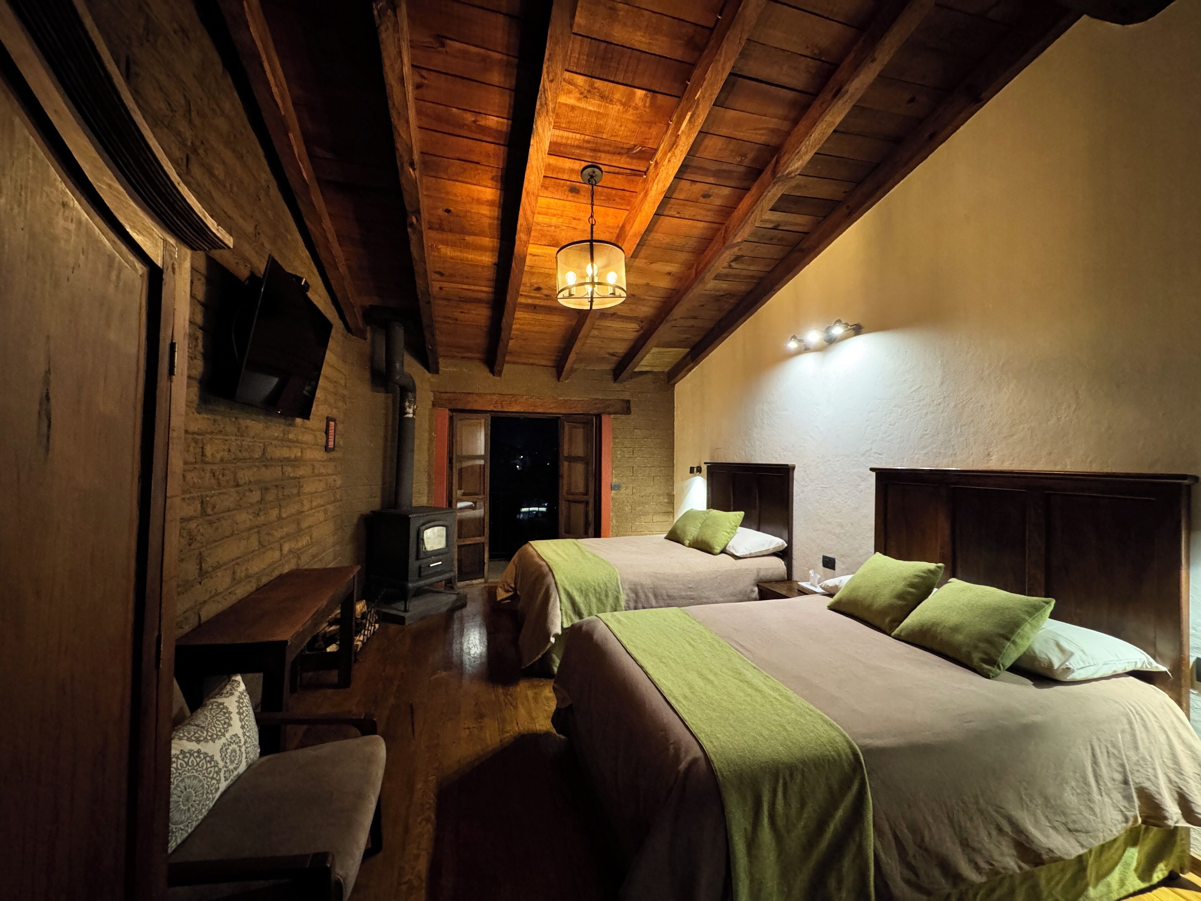 A cozy room with two beds, a wooden ceiling, a fireplace, and a TV mounted on the wall.