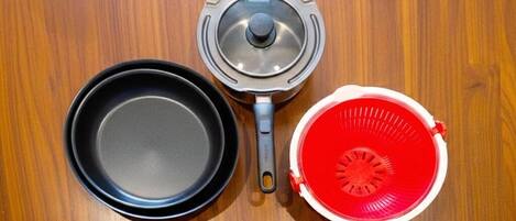 A wooden surface with three kitchen items: a black pan, a grey pot with a lid, and a red colander.
