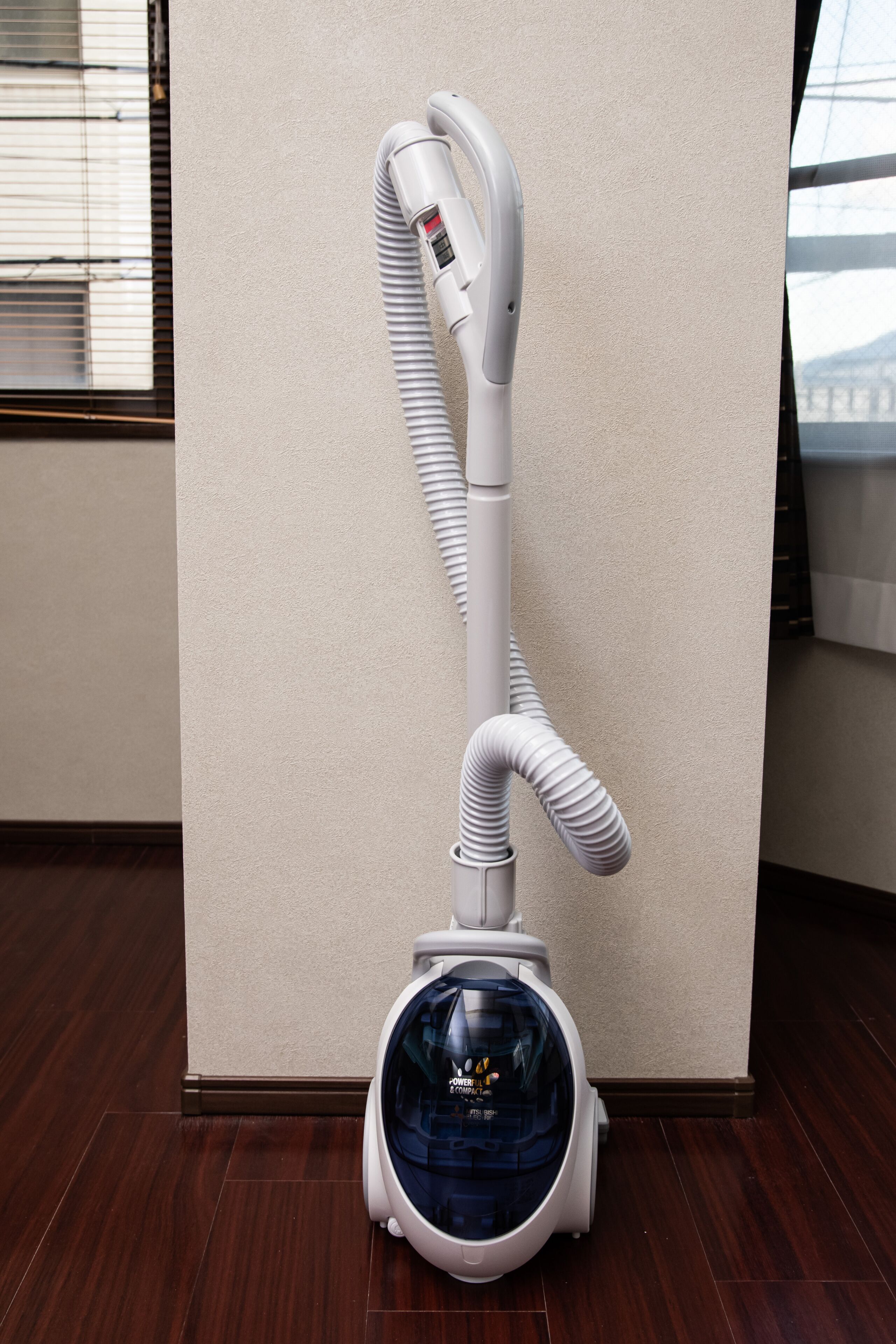 A vacuum cleaner with a flexible hose and a transparent dust container, positioned against a wall in a room with wooden flooring.